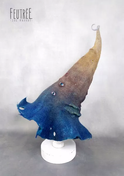 Chapeau fantastique de magicien en feutre artisanal, dégradé de couleur du bleu au beige, avec des éléments de détails tels que des yeux, des trous, des plis et griffures
