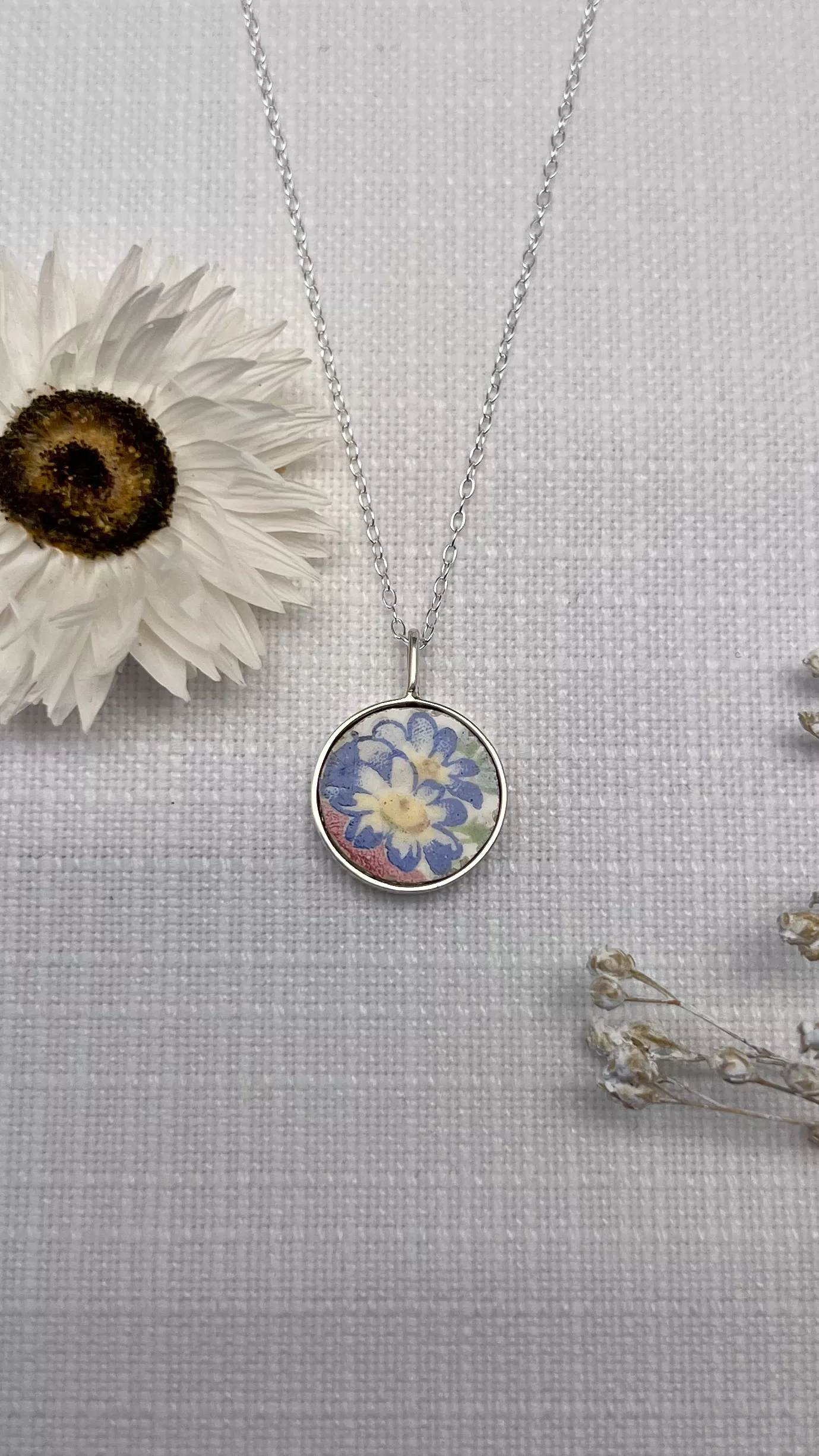 Pendentif rond en faïence ancienne à motif bleu et rose, monté sur chaîne argentée, présenté avec une fleur séchée en décoration.