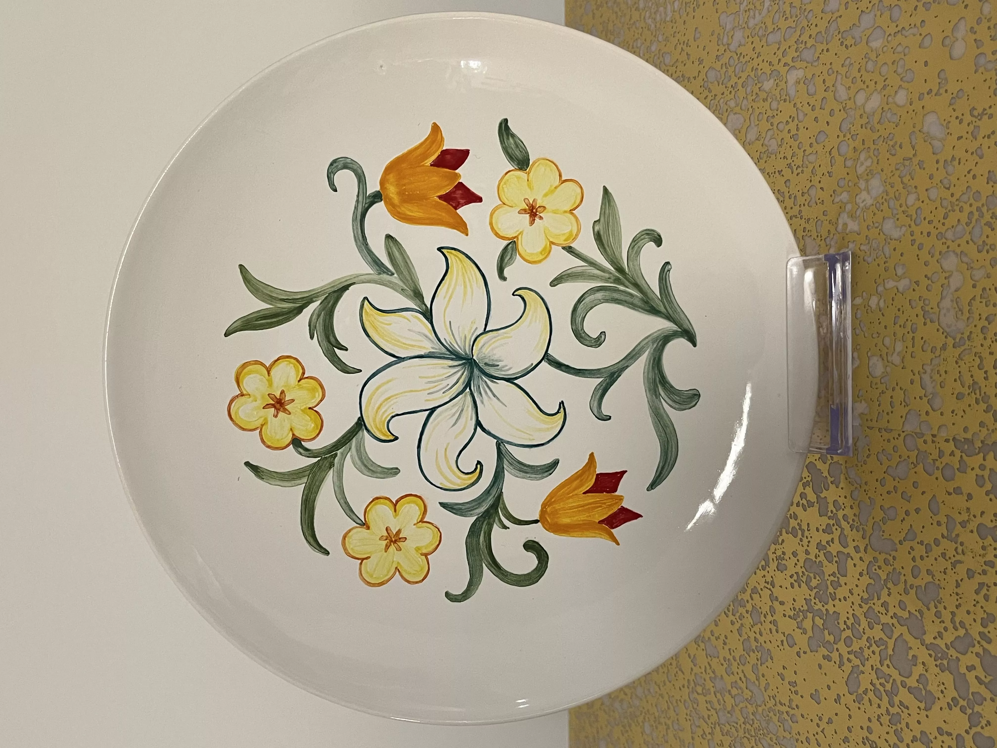Assiette en faïence de 21cm de diamètre, après cuisson. L'assiette présente un motif floral : des fleurs jaunes et orangées et des entrelacs de feuilles de différentes nuances de vert.