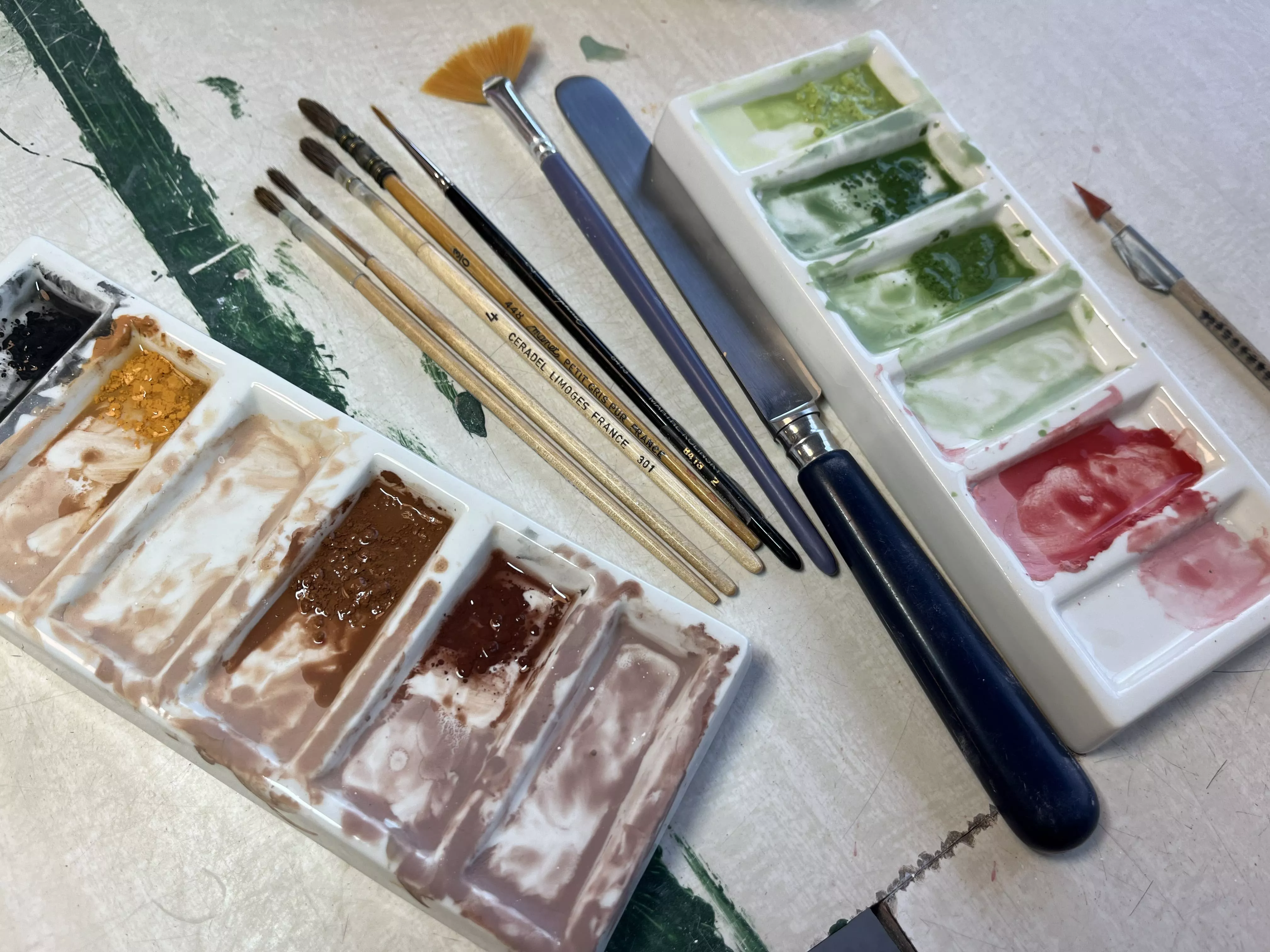 Photo du matériel nécessaire au décor sur céramique : différents pinceaux, des palettes avec des couleurs à peindre, un couteau pour mélanger la peinture en poudre à l'eau, et différents outils