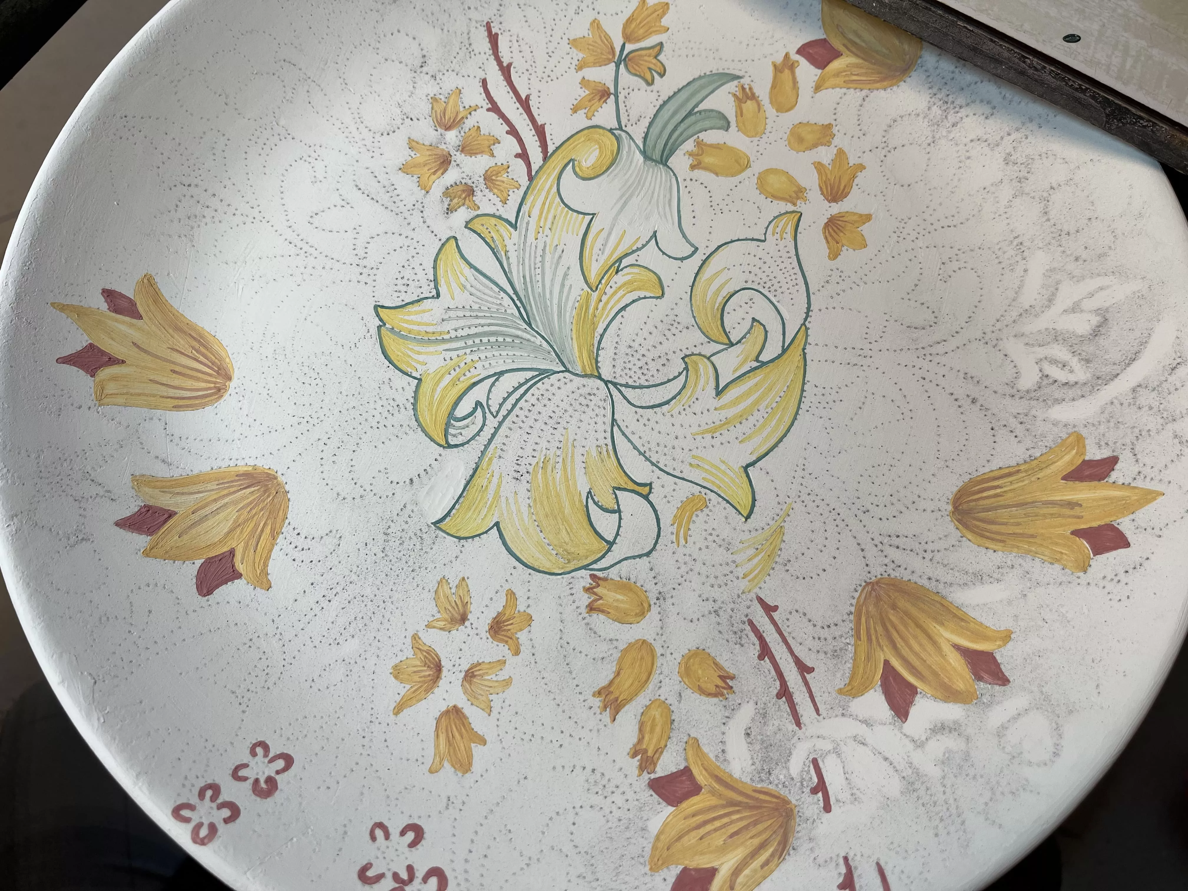 Photo d'une peinture de fleurs en cours de réalisation. La photo est prise du dessus comme si l'observateur était le peintre. On peut voir la trace du transfert du motif, il s'agit de fusain en poudre qui disparaîtra à la cuisson, seule la peinture restera. La particularité de la peinture sur émail est que l'ensemble des coups de pinceaux restera visible après la cuisson. 