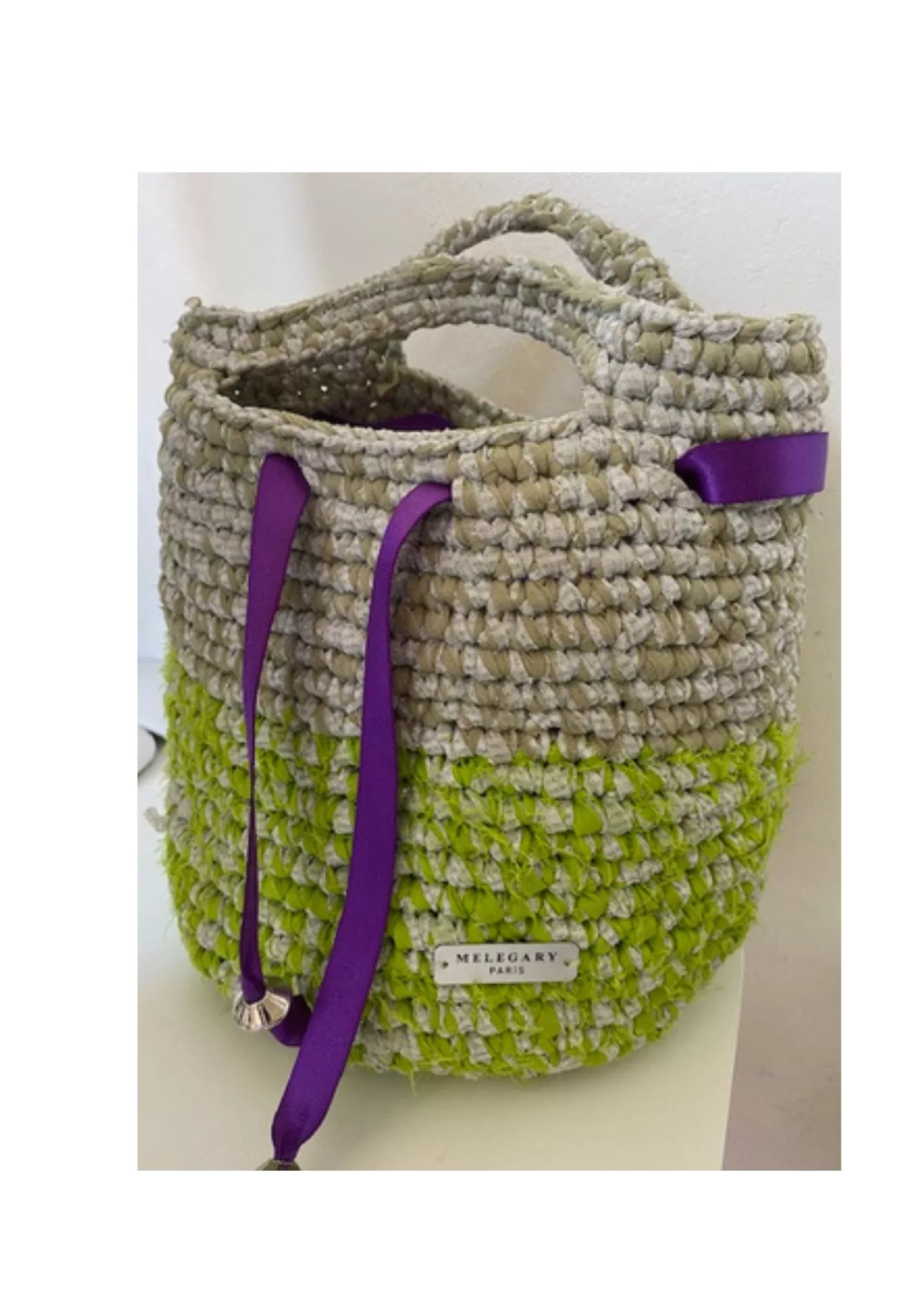 Ce sac à main est entièrement réalisé à la main à partir de bandes de tulle et de soie recyclées, travaillées au crochet pour créer une texture riche et volumineuse. Son design bicolore associe des nuances de vert anis et de beige naturel, rehaussées par d'élégants rubans violets qui apportent une touche contemporaine et raffinée