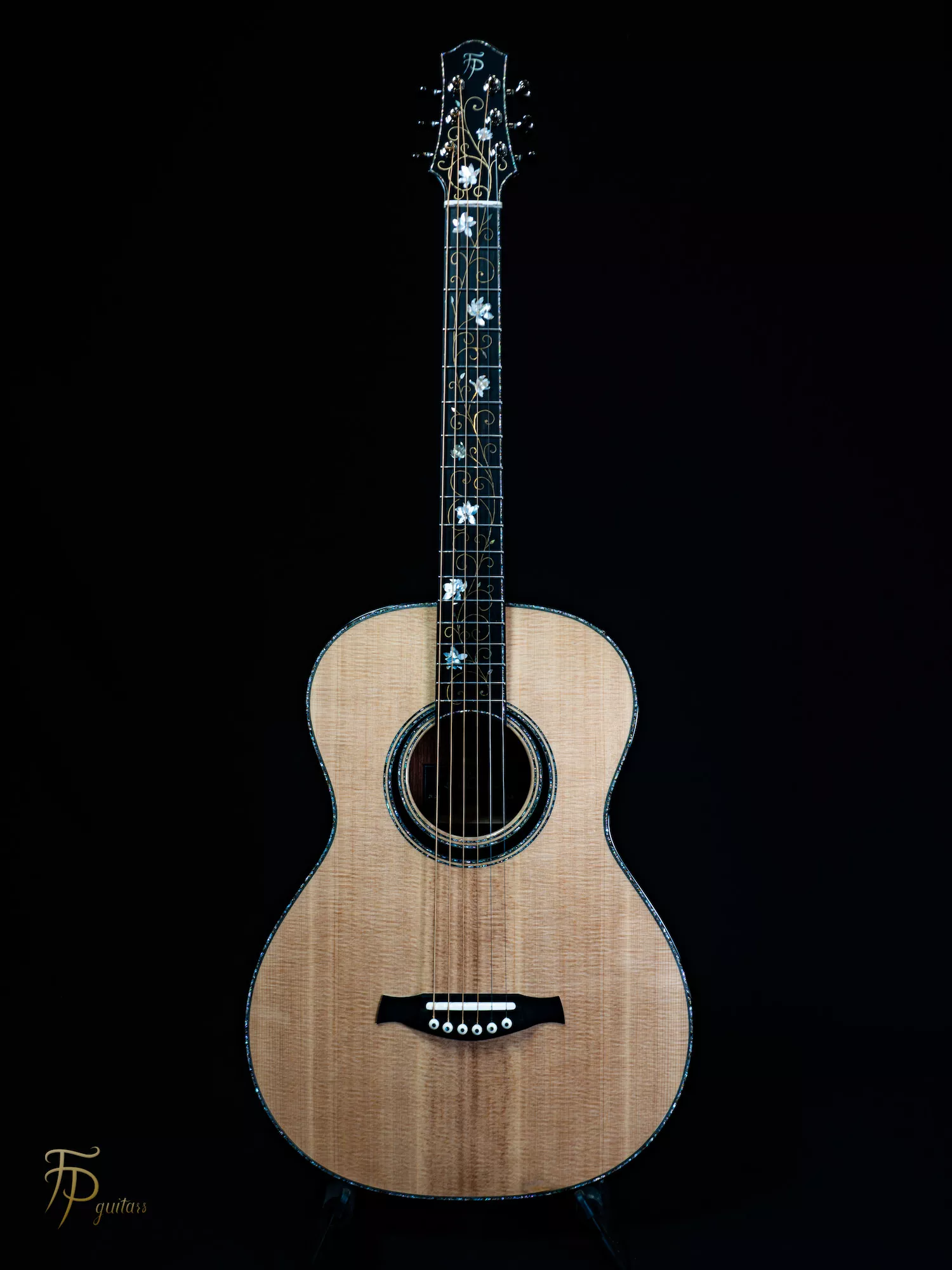 Guitare acoustique au format 00, entièrement réalisée à la main. Sa table en épicéa de Sitka est soulignée par des filets d’ébène et de nacre qui accompagnent le contour de la caisse et la rosace. Un décor floral en nacre et en laiton se déploie de la tête jusqu’à la touche, mêlant précision technique, finesse du trait et jeux de lumière. Cette ornementation a été pensée comme une composition continue, intégrée à l’architecture de l’instrument.