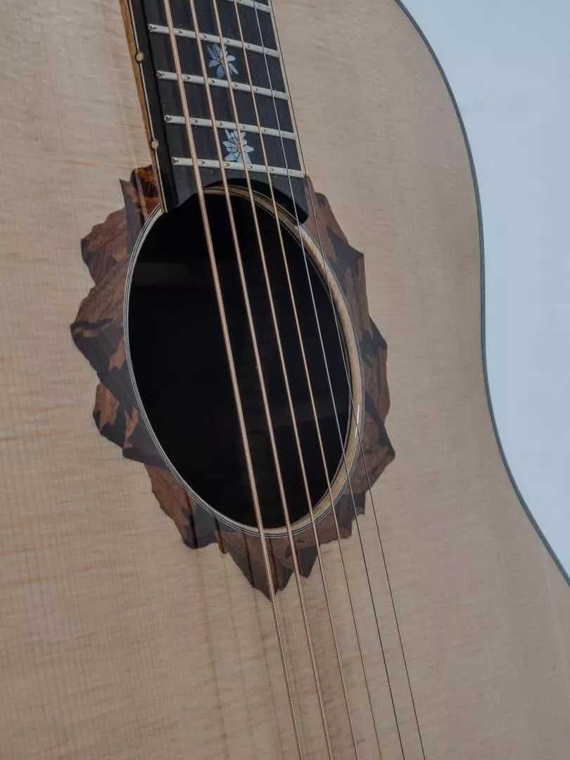 Détail d’une guitare Grand Auditorium mettant en valeur une rosace figurative en marqueterie, inspirée des reliefs montagneux. Les différentes essences de bois sont découpées puis assemblées pour créer un paysage aux nuances et aux profondeurs naturelles. Des repères de touche en forme d’edelweiss prolongent cette identité alpine et établissent une continuité visuelle entre la table et le manche.