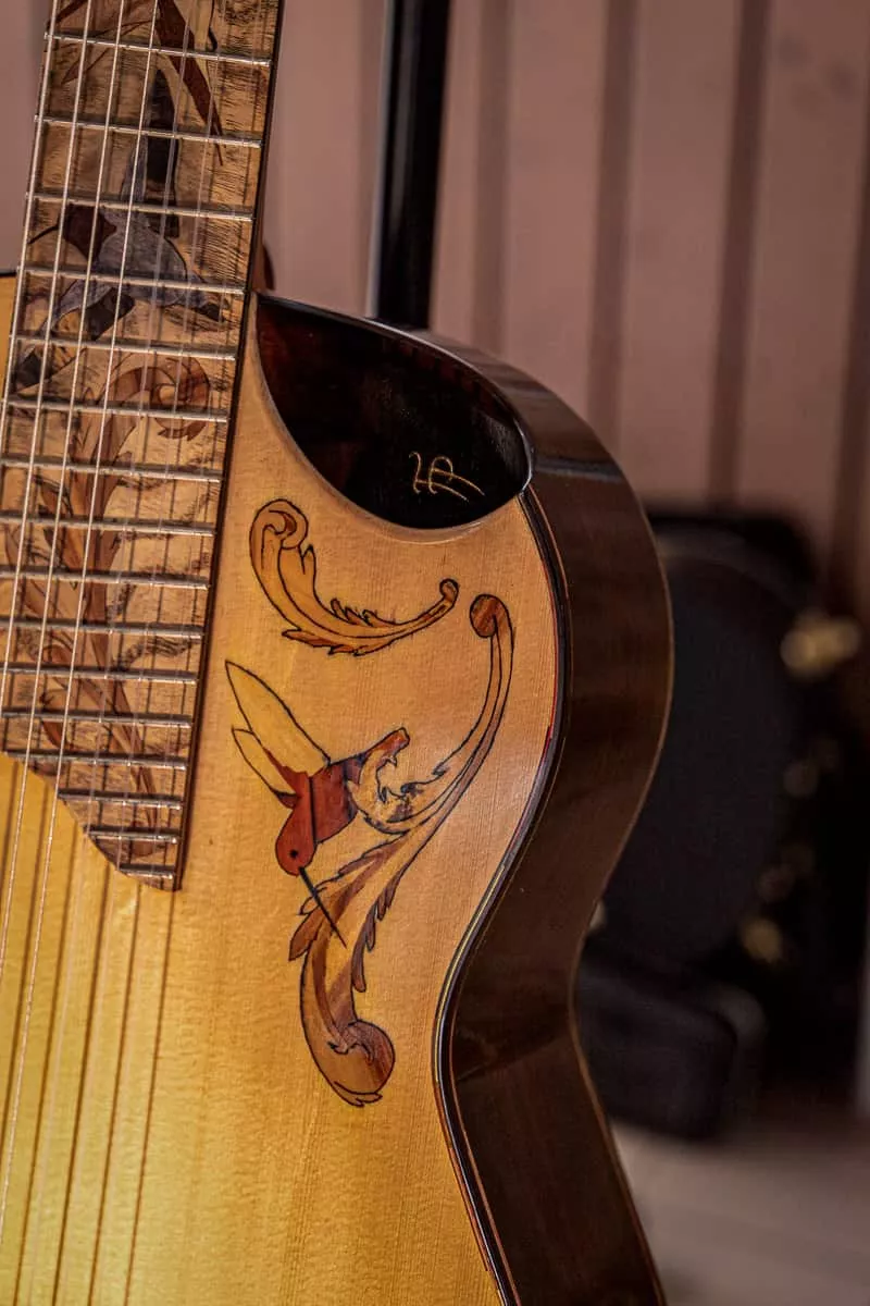 Détail d’une guitare classique à sept cordes dont le décor se développe de la touche jusqu’à la table. La touche est ornée d’une marqueterie représentant plusieurs oiseaux, composée de pièces de bois aux teintes contrastées. Sur la table, une incrustation figurative prolonge ce motif par un oiseau stylisé et des formes végétales. Chaque élément est découpé, ajusté et intégré à la main pour former une composition continue autour de l’instrument.