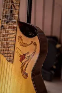 Détail d’une guitare classique à sept cordes dont le décor se développe de la touche jusqu’à la table. La touche est ornée d’une marqueterie représentant plusieurs oiseaux, composée de pièces de bois aux teintes contrastées. Sur la table, une incrustation figurative prolonge ce motif par un oiseau stylisé et des formes végétales. Chaque élément est découpé, ajusté et intégré à la main pour former une composition continue autour de l’instrument.